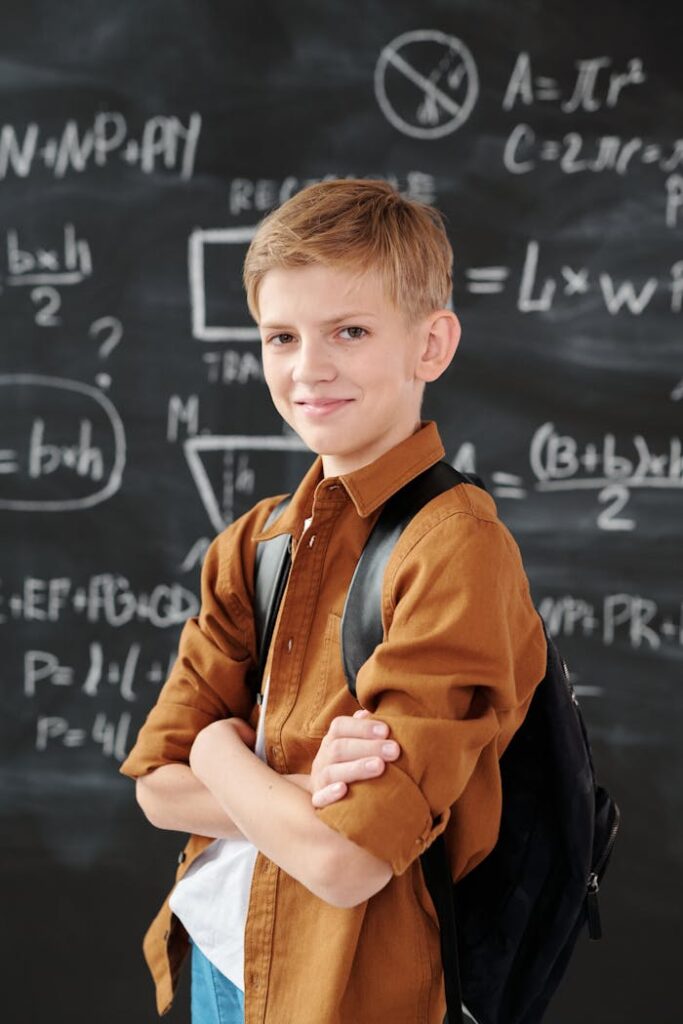 Smiling elementary student standing in classroom with math equations on blackboard.